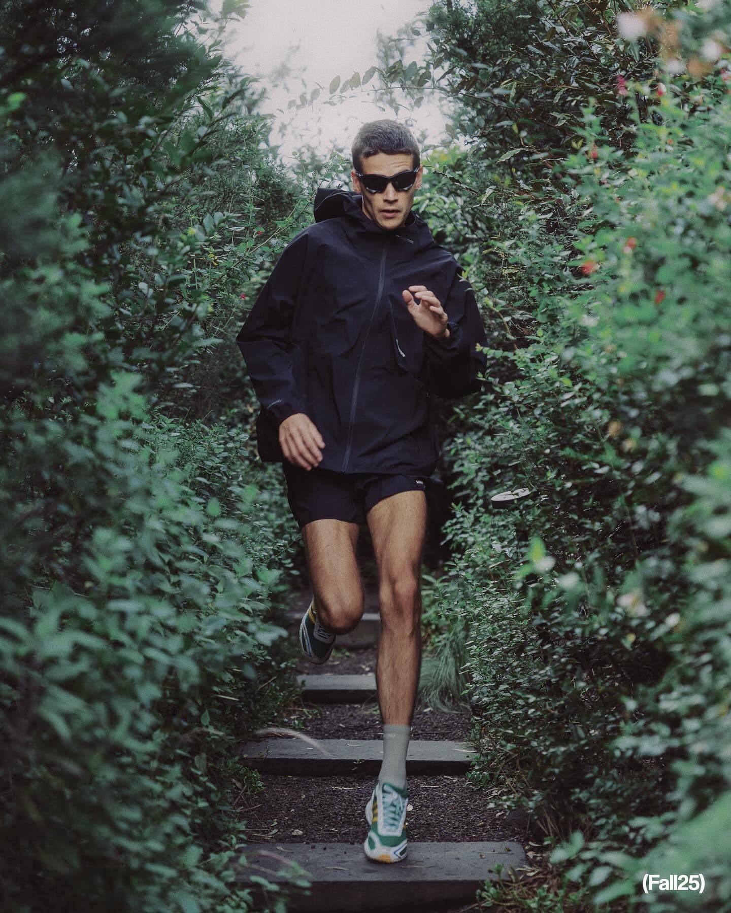 A man in a black jacket and sunglasses runs on a green, forested path.