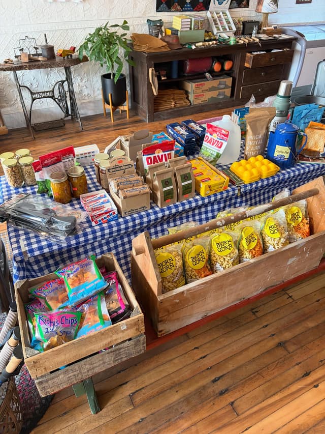 A general store display with packaged food and snacks on a blue and white checkered tablecloth, against a background of rustic wooden furniture and decor.