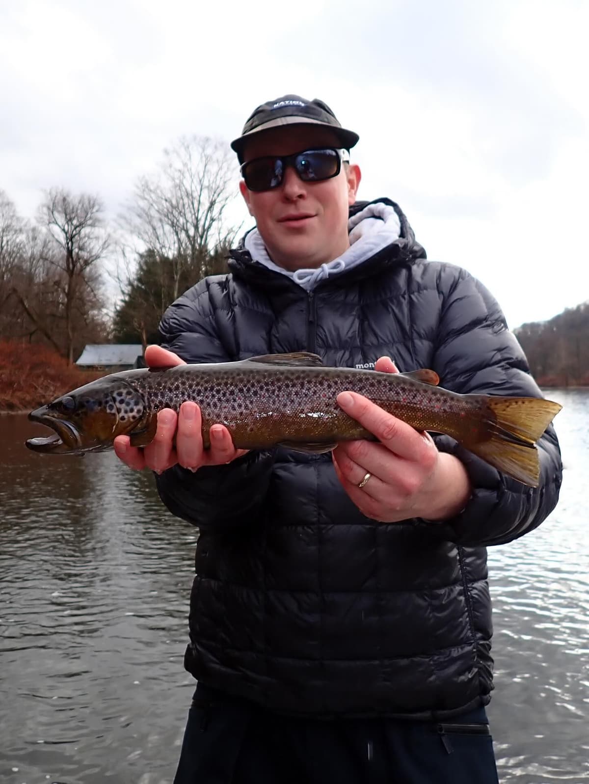 A man in a black jacket and sunglasses holds a large brown trout over a river.