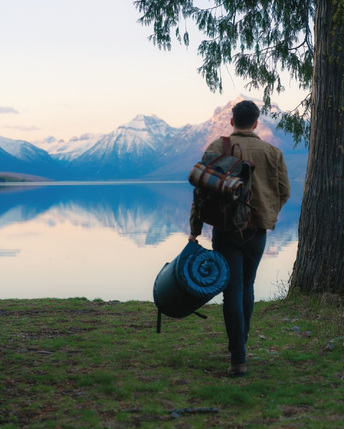 A person with a backpack and sleeping roll walks towards a lake reflecting snow-capped mountains.