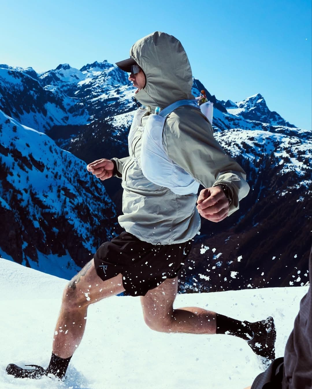 A man in a hooded jacket and shorts runs through snow, kicking it up, with snow-covered mountains in the background under a clear blue sky.