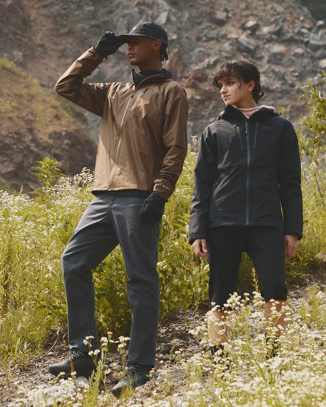 A man and woman in hiking jackets stand among wildflowers against a rocky backdrop.