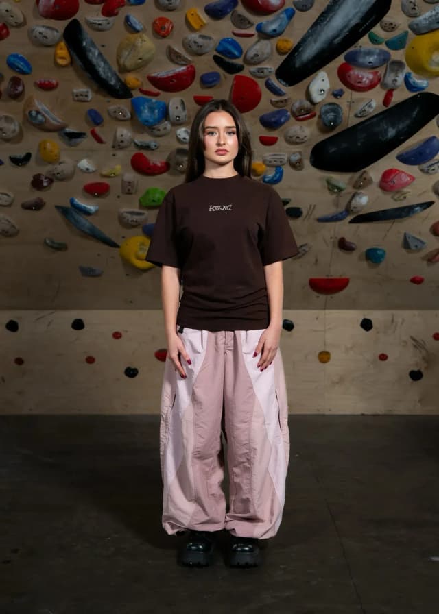 A young woman in a brown t-shirt and pink paneled cargo pants stands against a climbing wall.