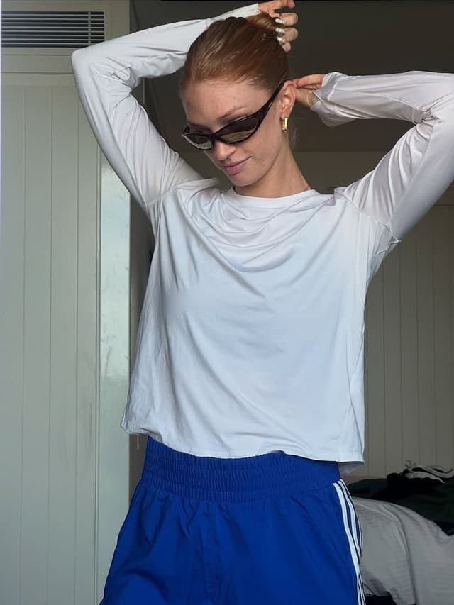 Red-haired woman in a white long-sleeve top and blue shorts adjusts her bun while wearing sunglasses.