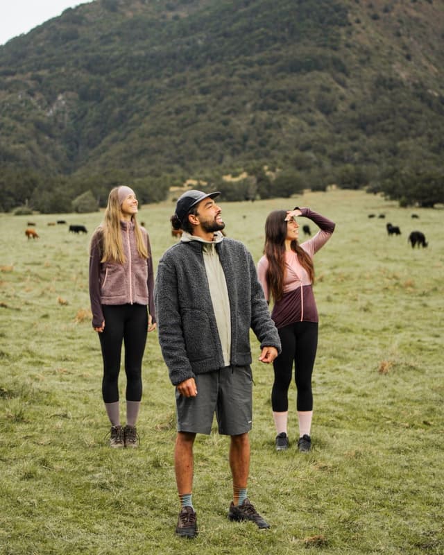 Three people in outdoor clothing stand in a green field, looking up and smiling, with mountains and grazing cows in the background.