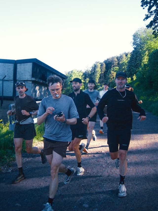 Group of men running on an outdoor trail. The lead runner wears glasses, a mouthguard, and holds a small device.