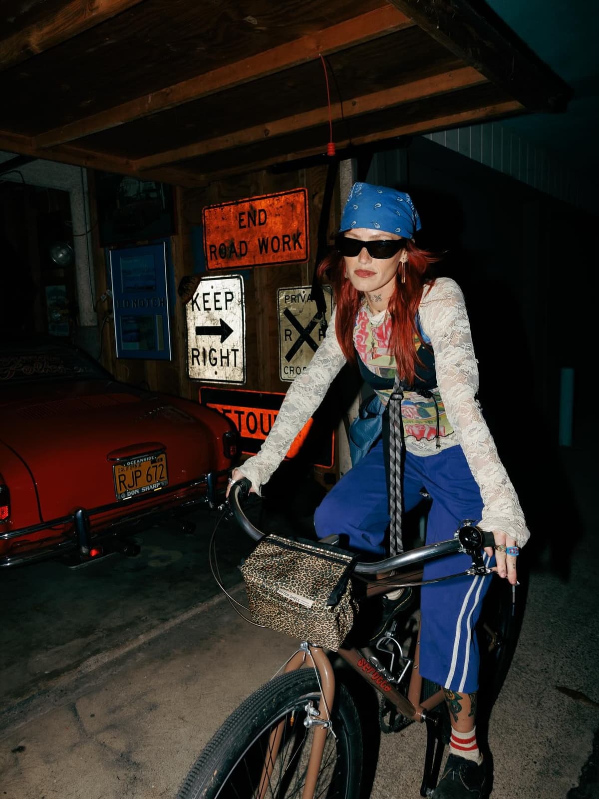A person with red hair, a blue bandana, and sunglasses rides a bicycle in a dimly lit space with road signs and a vintage red car in the background.