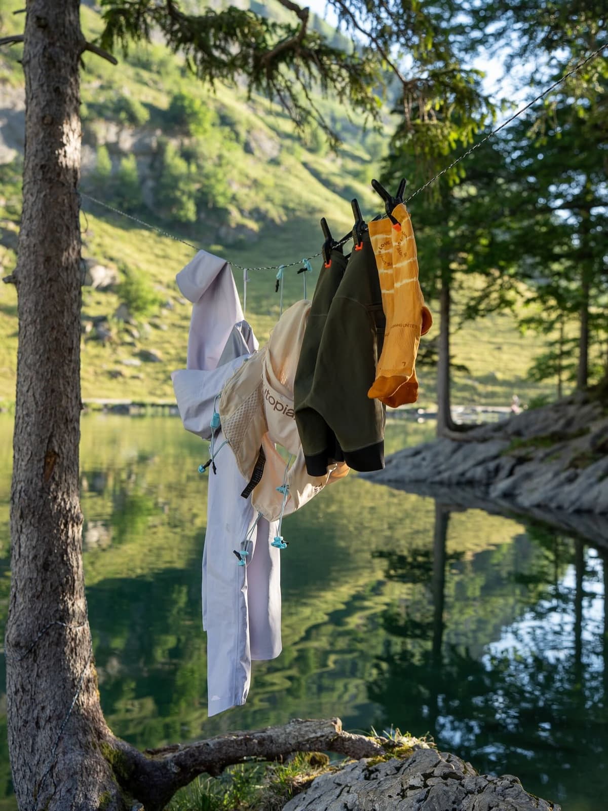 Clothes drying on a line strung between trees beside a calm lake with green mountains in the background.