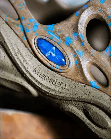 Close-up of a tan Merrell shoe or sandal speckled with blue, featuring the brand name embossed and a blue oval emblem.