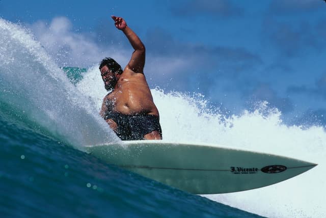 A large, shirtless, bearded man surfs a wave.