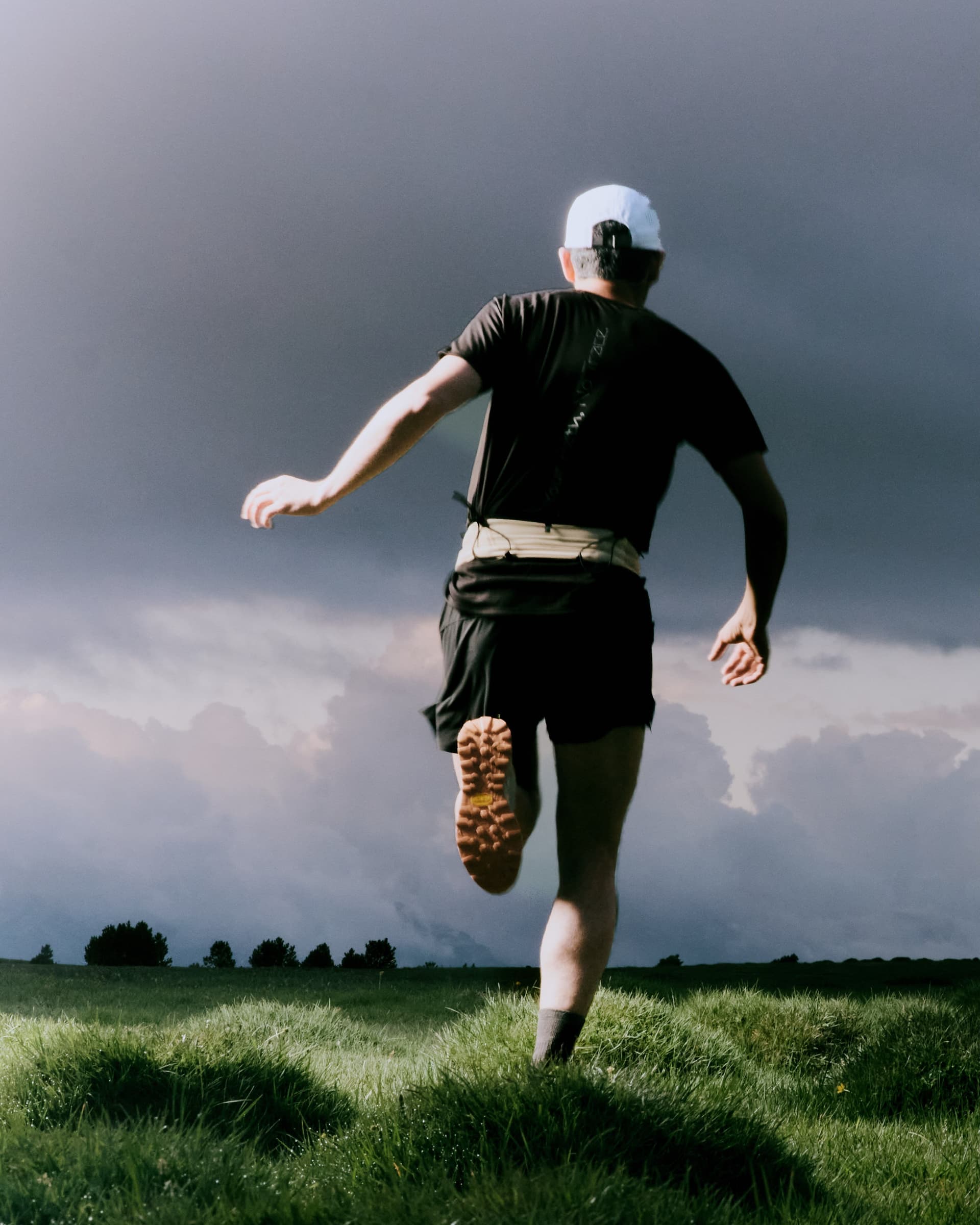 Runner in dark gear and a white cap from behind, mid-stride on a grassy field under a stormy sky.
