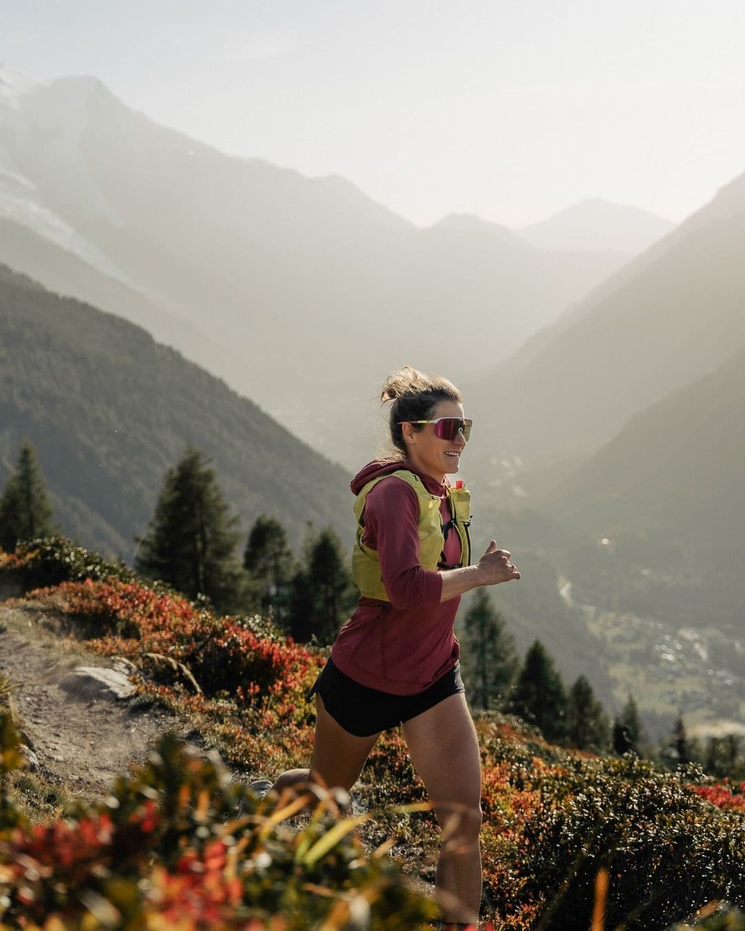 A woman trail runs on a mountain path with a hazy valley and peaks in the background, surrounded by autumn foliage.
