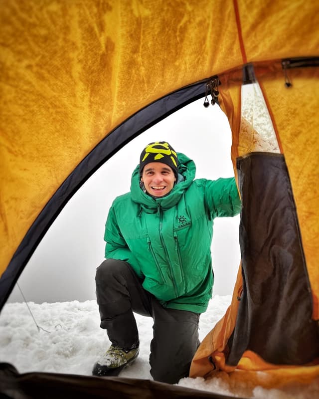 A smiling man in a green jacket and beanie kneels in the snow, looking out from an orange tent.