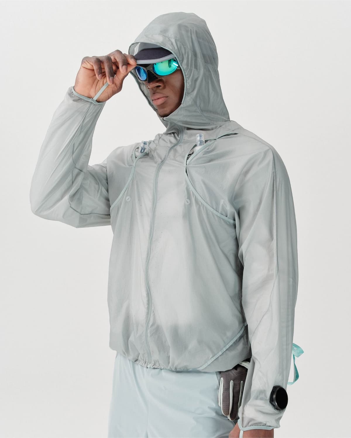 A man wears a translucent gray hooded jacket, cap, and mirrored sunglasses, adjusting his cap.