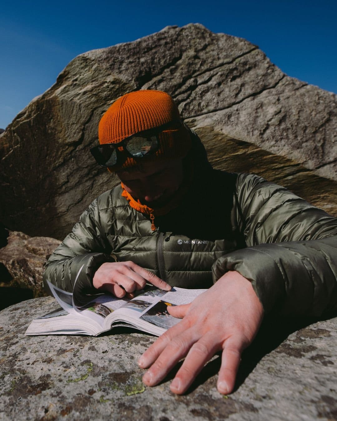 A person in an orange beanie and green jacket points to a page in an open book while sitting on a large rock outdoors.
