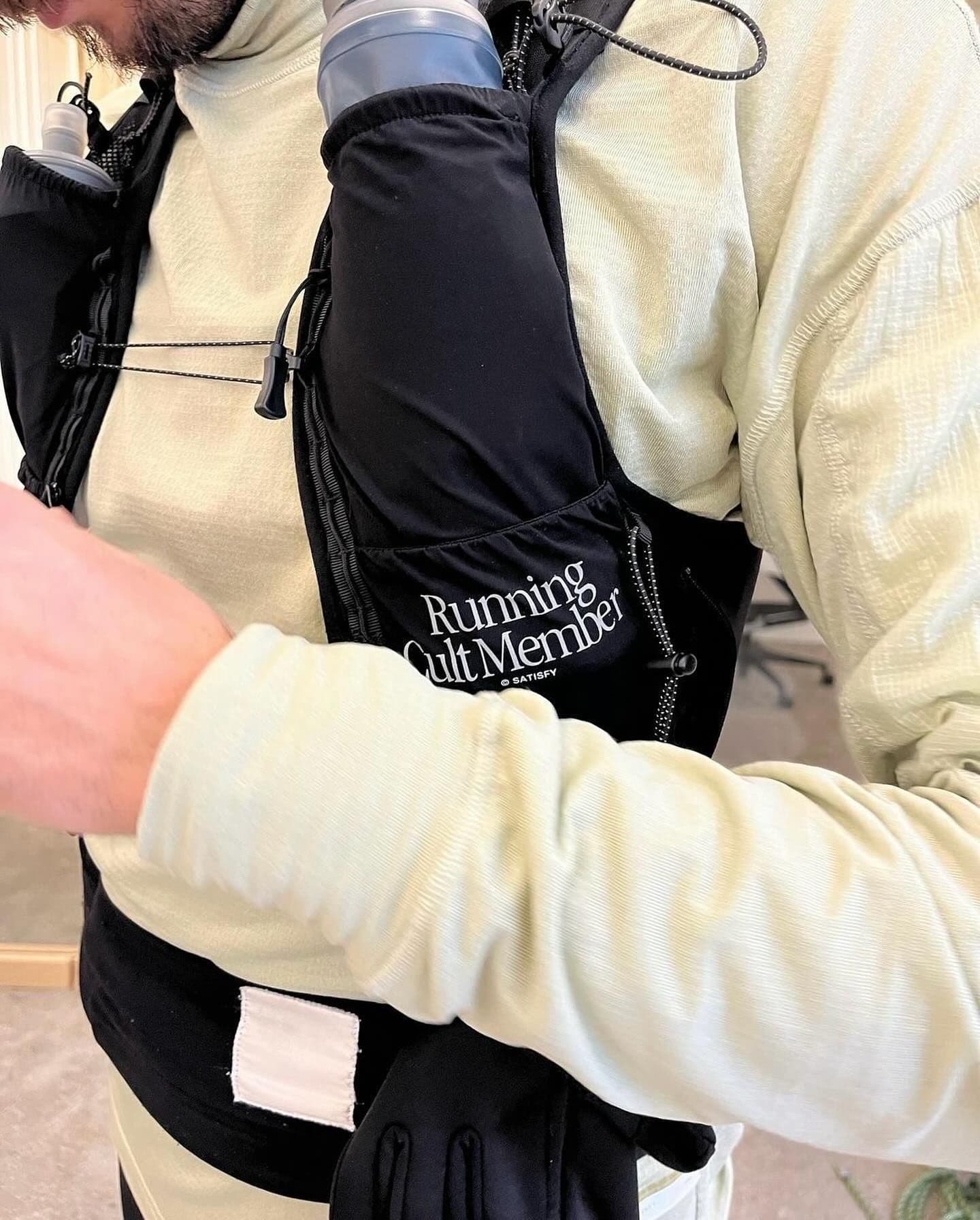 A person wearing a black running vest with two water bottles over a light green long-sleeved shirt, with "Running Cult Member" visible on the vest.