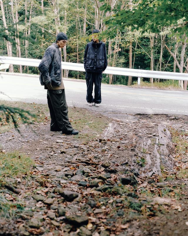 Two people in outdoor wear stand on a rocky path next to a road, with a forest behind them.