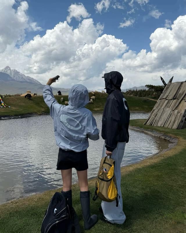 Two people stand by a lake, one holding up a phone, with mountains, yaks, and sheep in the scenic background.