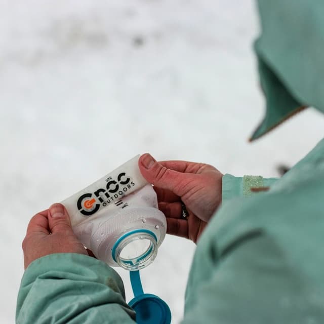 Hands holding a "Cnoc Outdoors" soft water bottle with a blue cap in a snowy environment.