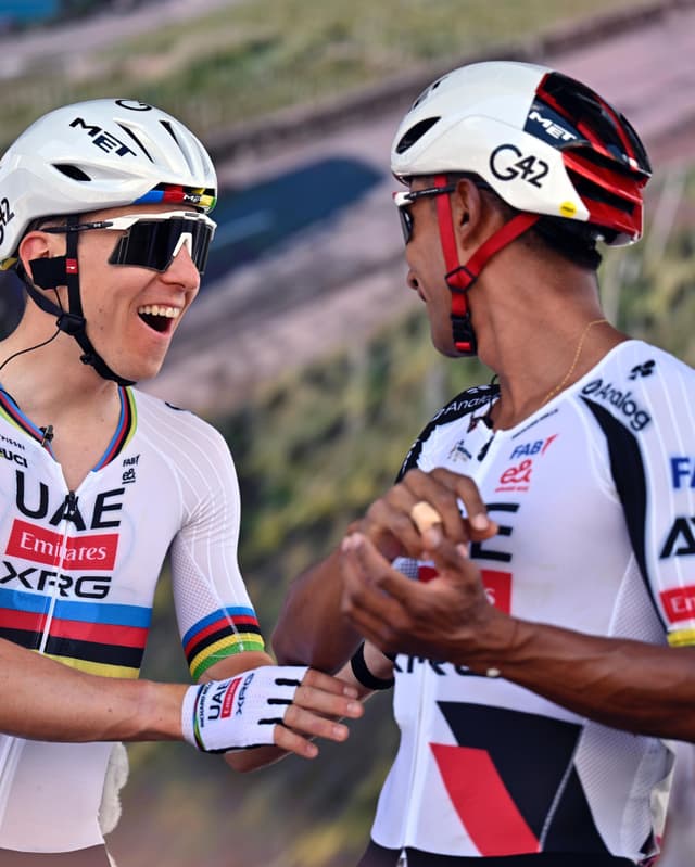 Two male cyclists, one with a rainbow-striped sleeve, shake hands and smile.