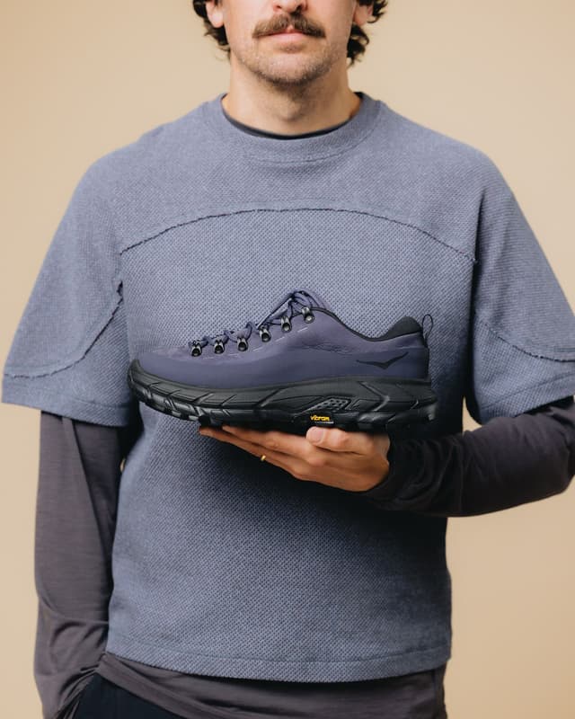 Man with a mustache holding a dark blue Hoka hiking shoe with a black Vibram sole.