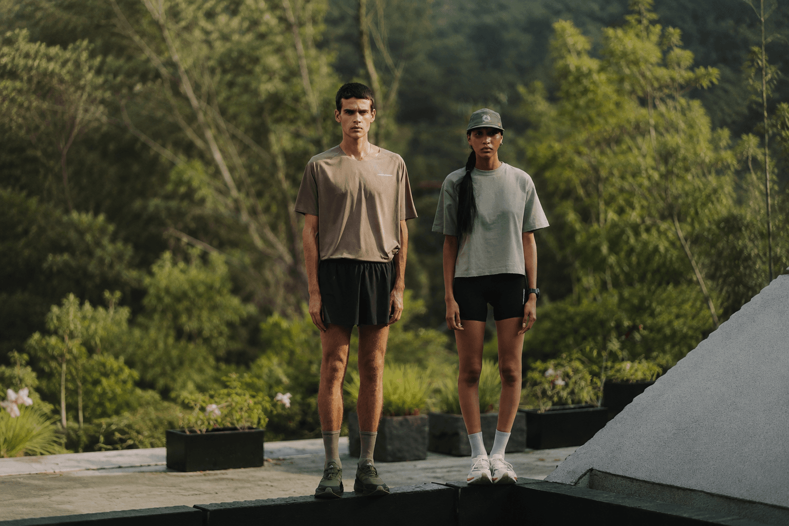 A man in a brown tee and black shorts and a woman in a sage tee, black shorts, and a green cap stand outdoors among green trees.