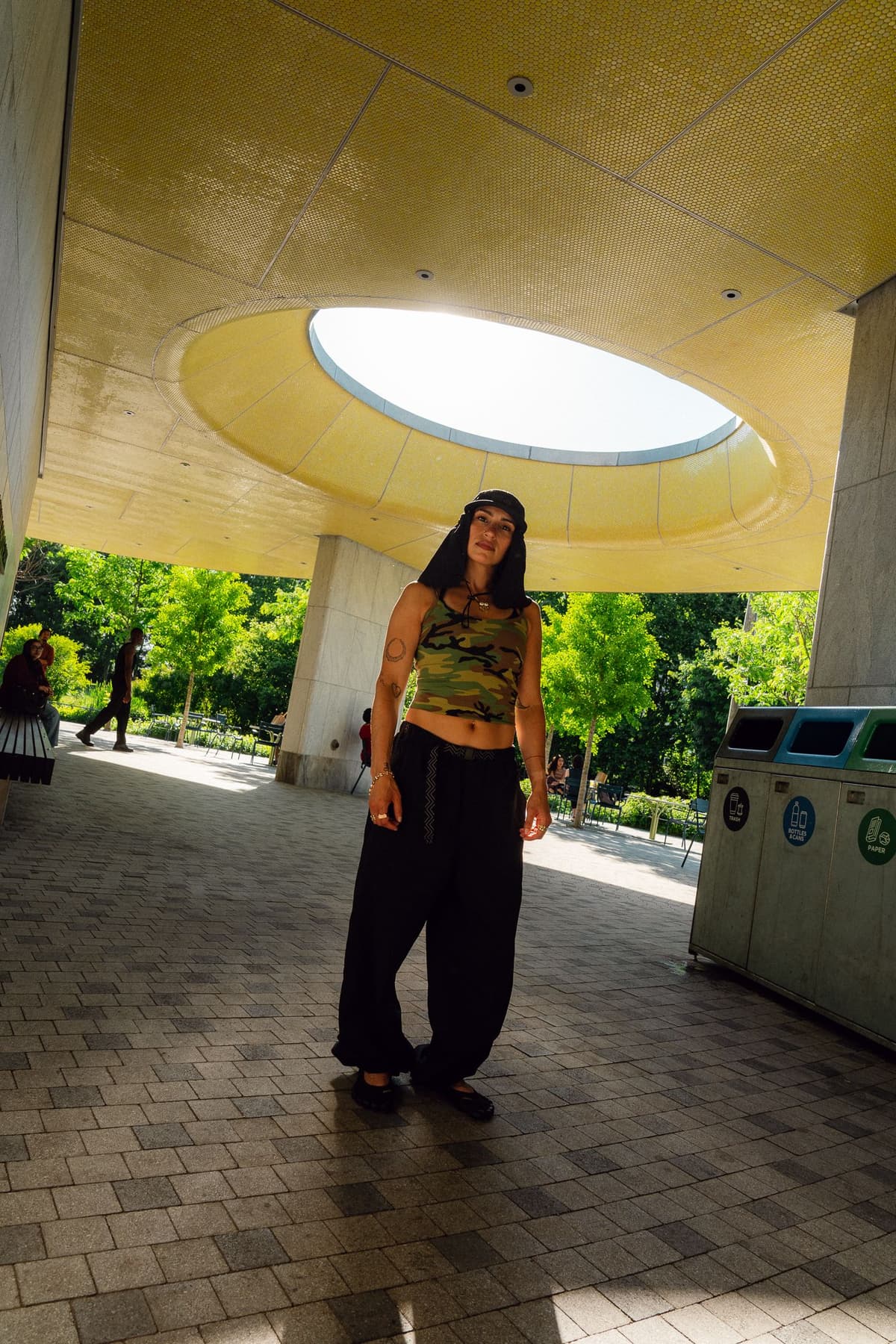 A woman in a camouflage crop top and black baggy pants stands under a modern structure with a circular skylight.