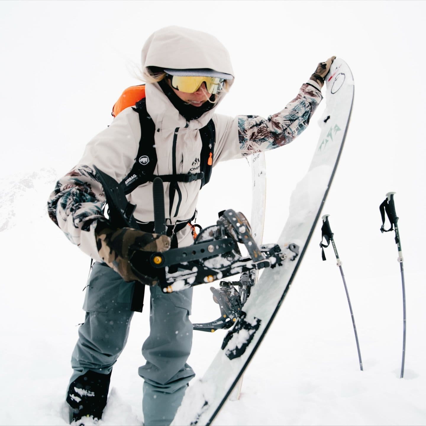 A person in winter gear holds a snowboard in a snowy landscape with trekking poles.