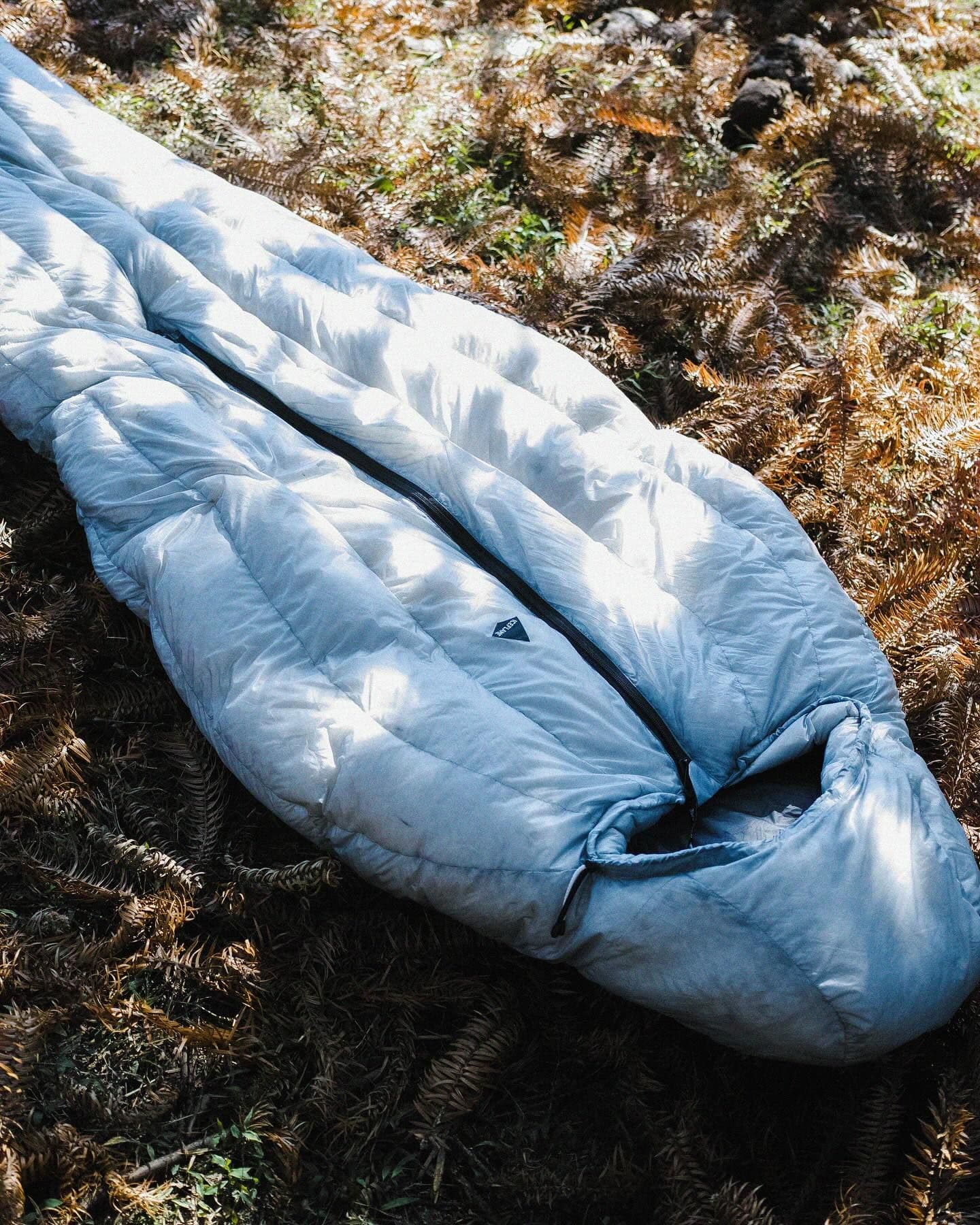 A light-colored sleeping bag lies partially unzipped on dried ferns.