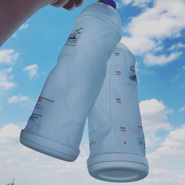 Two white collapsible water bottles held up against a blue sky with clouds.