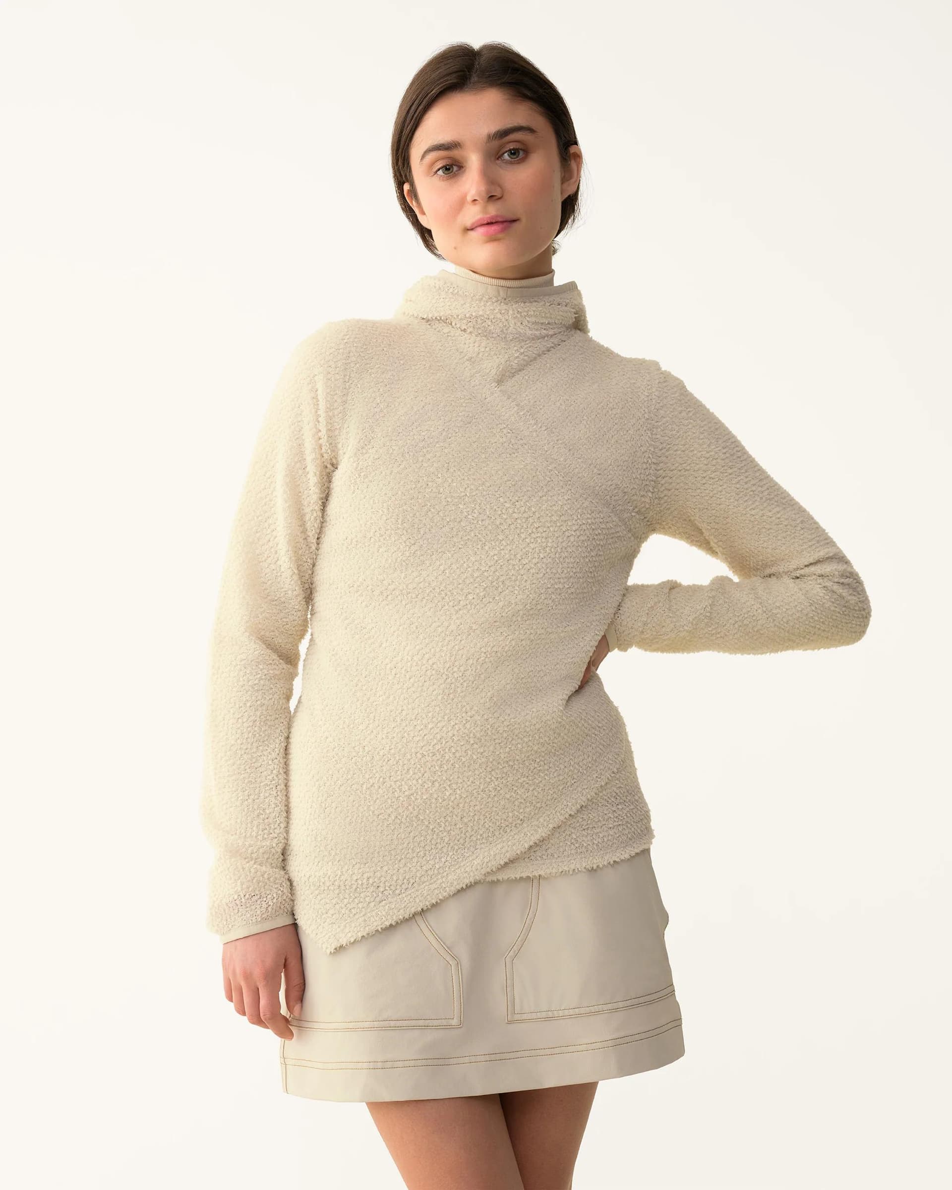 A woman models a cream textured asymmetrical wrap sweater and a light beige skirt with visible stitching.
