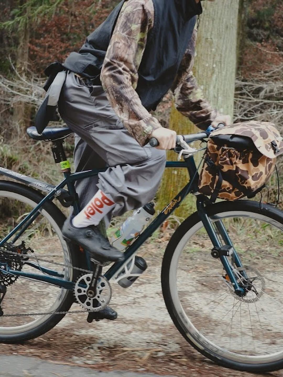 A person in a camouflage long-sleeve shirt and dark vest rides a bicycle with a camouflage front bag on a dirt path in a forest.