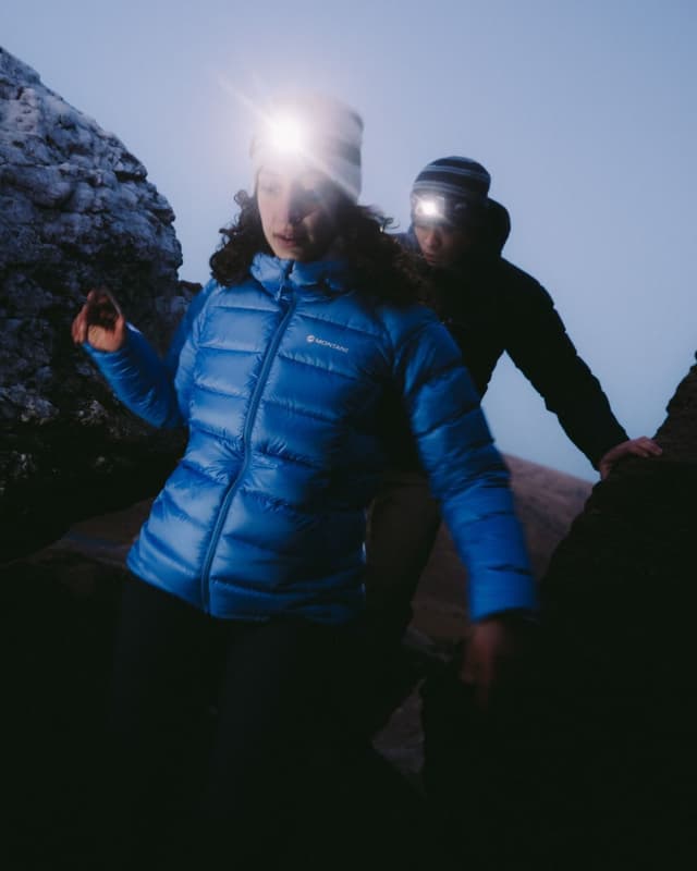 Two hikers wearing bright headlamps navigate dark, rocky terrain.