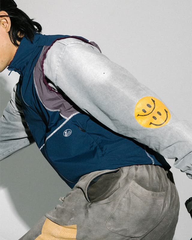A person wears a blue vest over a gray long-sleeved shirt with two yellow smiley faces on the left arm, and gray sweatpants.