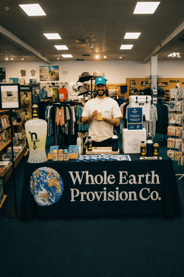 A smiling man in a retail store stands behind a "Whole Earth Provision Co." table displaying a Nuun drink dispenser and other products.