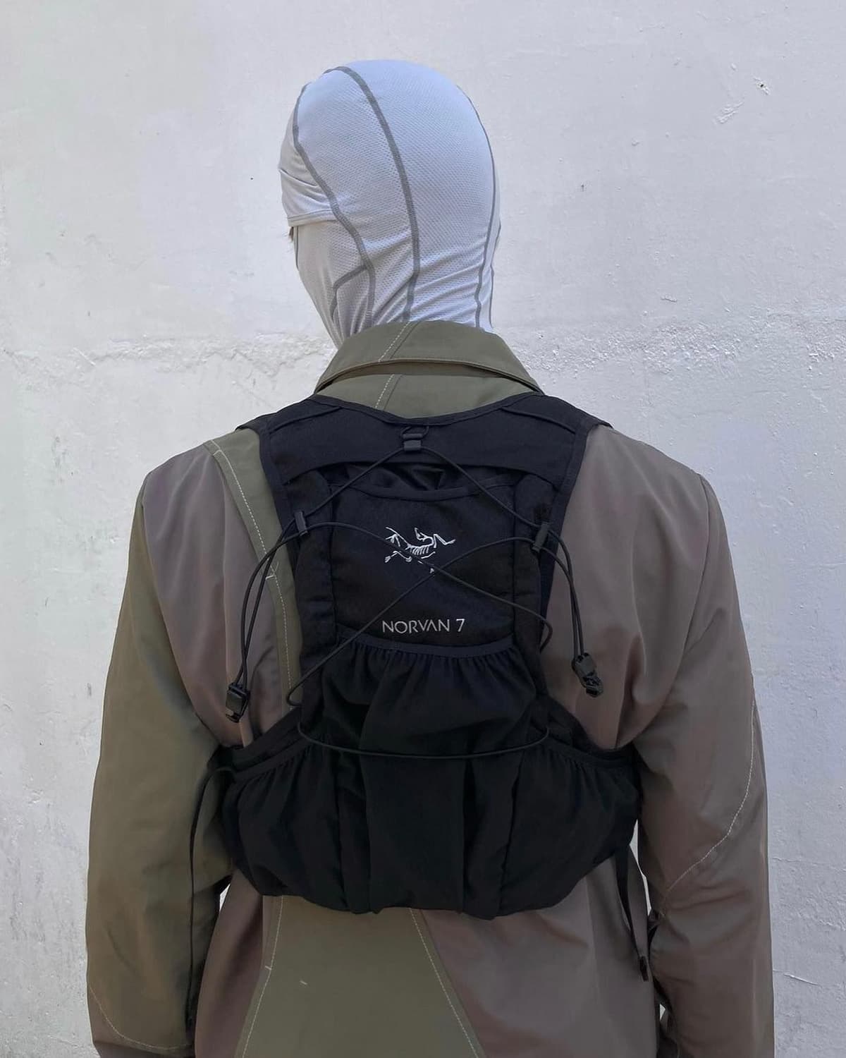 Back view of a person wearing a light gray balaclava, a brown jacket, and a black Arc'teryx Norvan 7 running vest.
