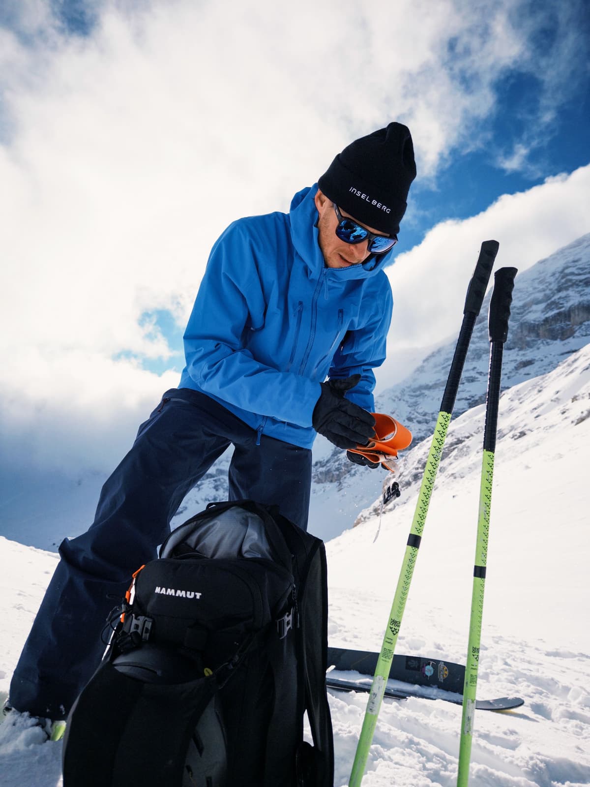 A man in a blue jacket and black hat adjusts orange ski equipment on a snowy slope with a backpack and ski poles nearby.