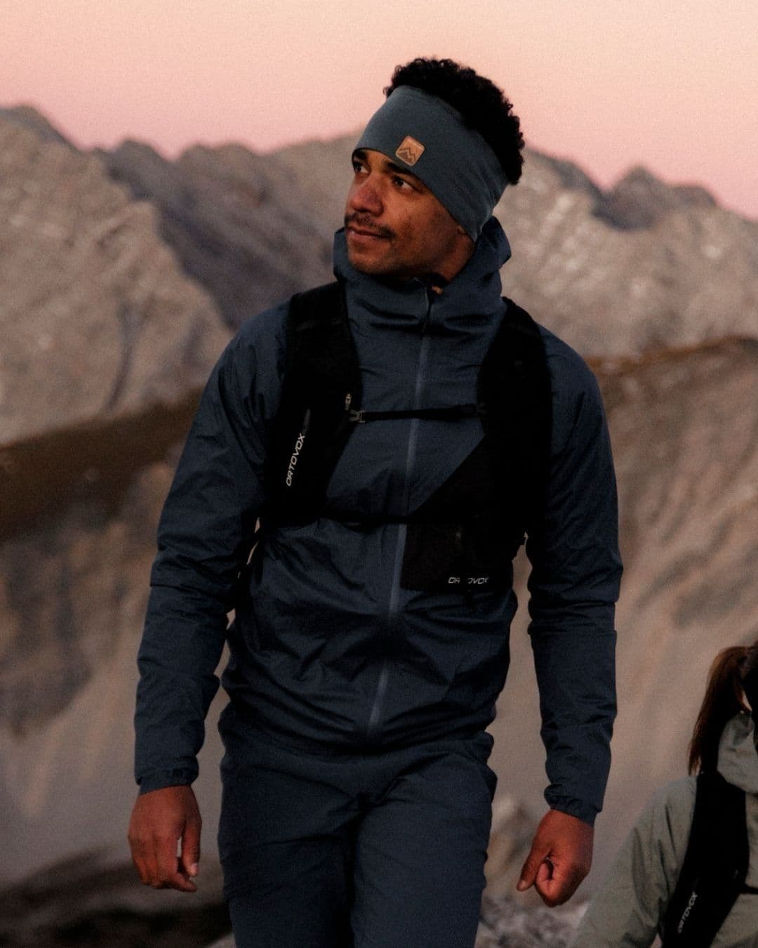 A man wearing a headband and dark athletic jacket hikes in a rocky mountain landscape.