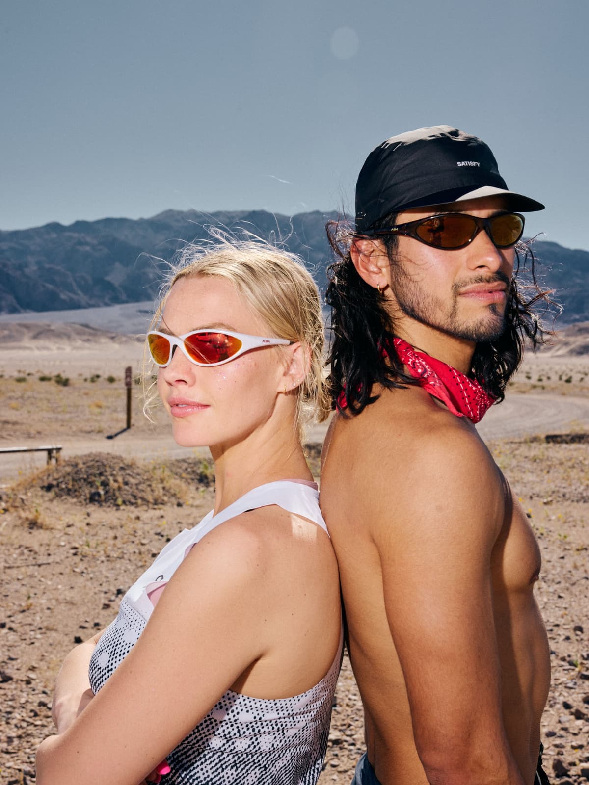 A blonde woman and a dark-haired man in athletic wear stand back-to-back in a desert landscape.