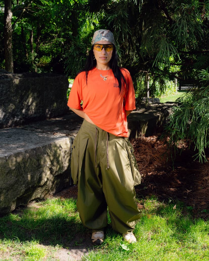 A woman in an orange t-shirt, olive green baggy pants, patterned headscarf, and yellow sunglasses stands in a leafy park.