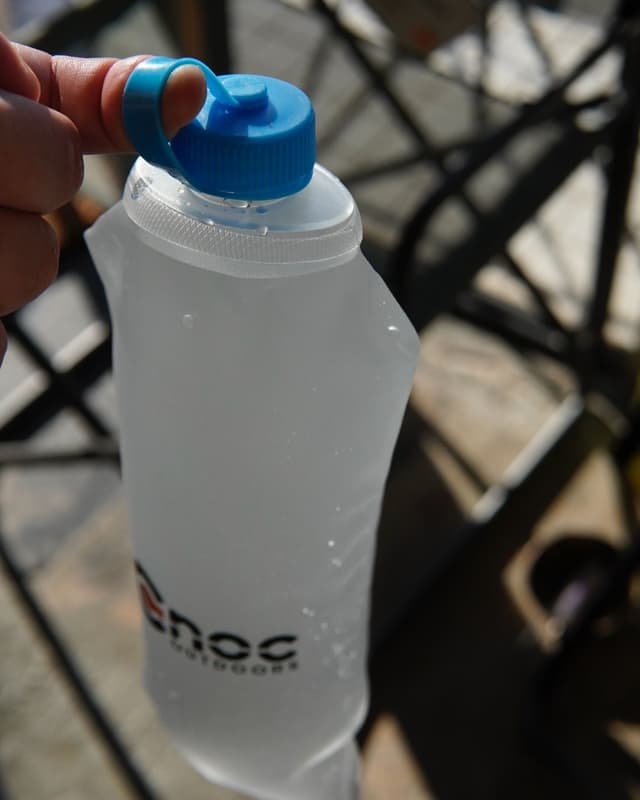 A hand holds a translucent flexible "Enoc Outdoors" water bottle with a blue cap, covered in water droplets.