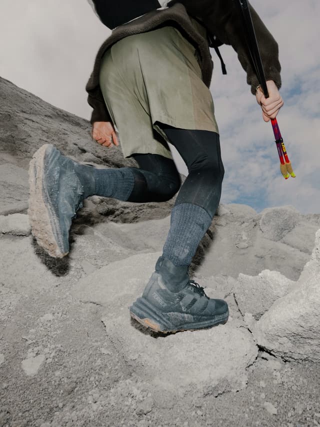 A person's lower body, dressed in shorts, leggings, and dark trail shoes, steps up a dusty, rocky incline while holding colorful trekking poles.