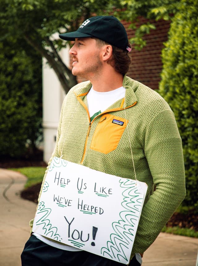 A man in a black cap and green Patagonia fleece looks left, holding a sign that says "Help Us Like WE'VE HELPED You!".