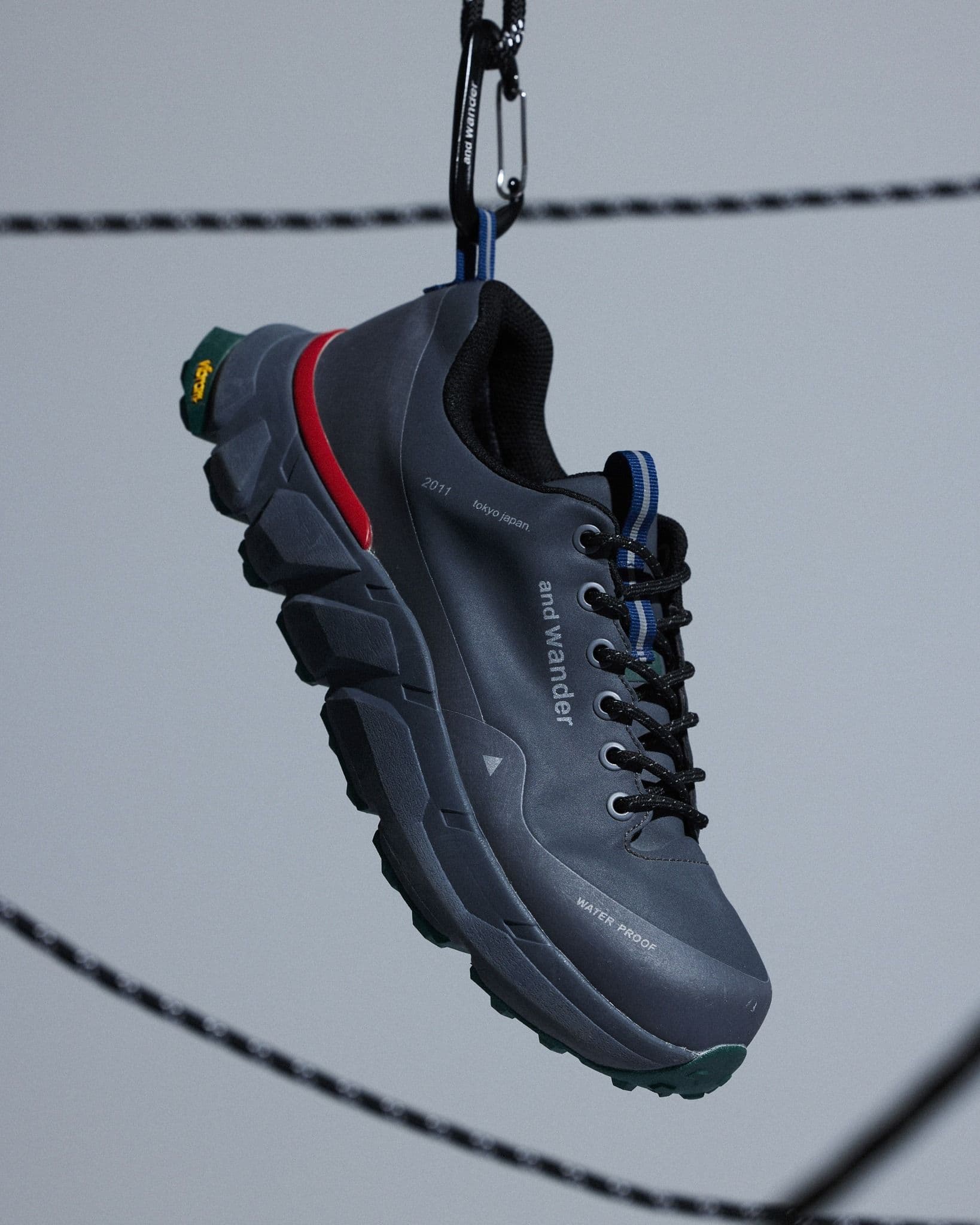 A dark gray And Wander hiking shoe with a red stripe and green Vibram sole, suspended by a carabiner.