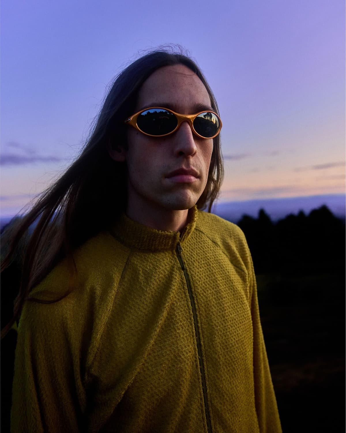 A person with long hair and yellow oval sunglasses wears a mustard jacket against a purple twilight sky.