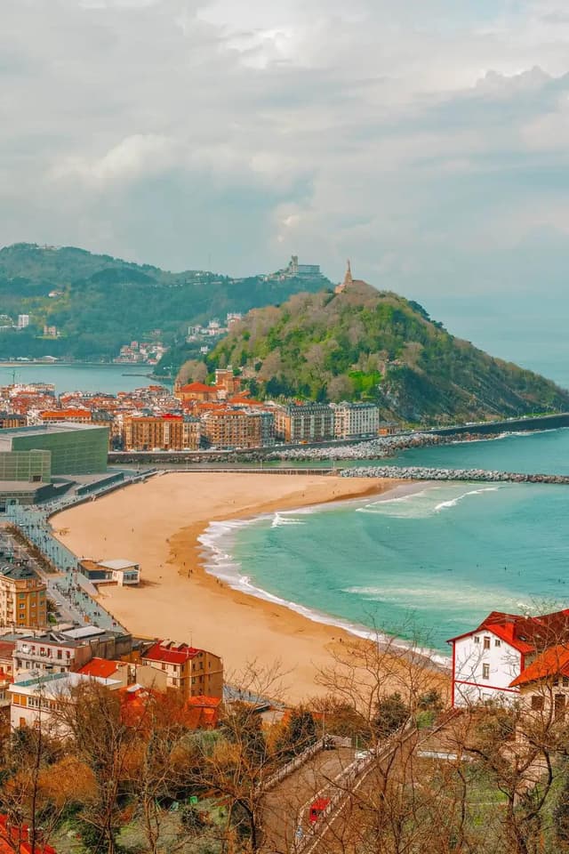 Aerial view of a coastal city with a crescent sandy beach, turquoise bay, urban buildings, and a forested hill topped with a statue and fortress.