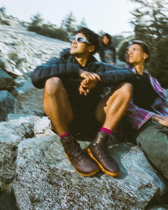 Three people sitting on rocks in a rugged outdoor landscape. The foreground man wears white sunglasses, a dark jacket, and brown boots with pink socks.