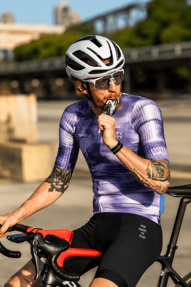 A male cyclist in a purple jersey and helmet eats an energy gel while sitting on his bike outdoors.