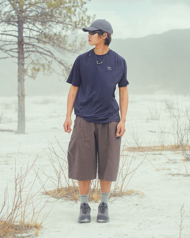 A young man in a cap, sunglasses, blue t-shirt, and brown shorts stands in a barren, light-colored landscape with a sparse tree.