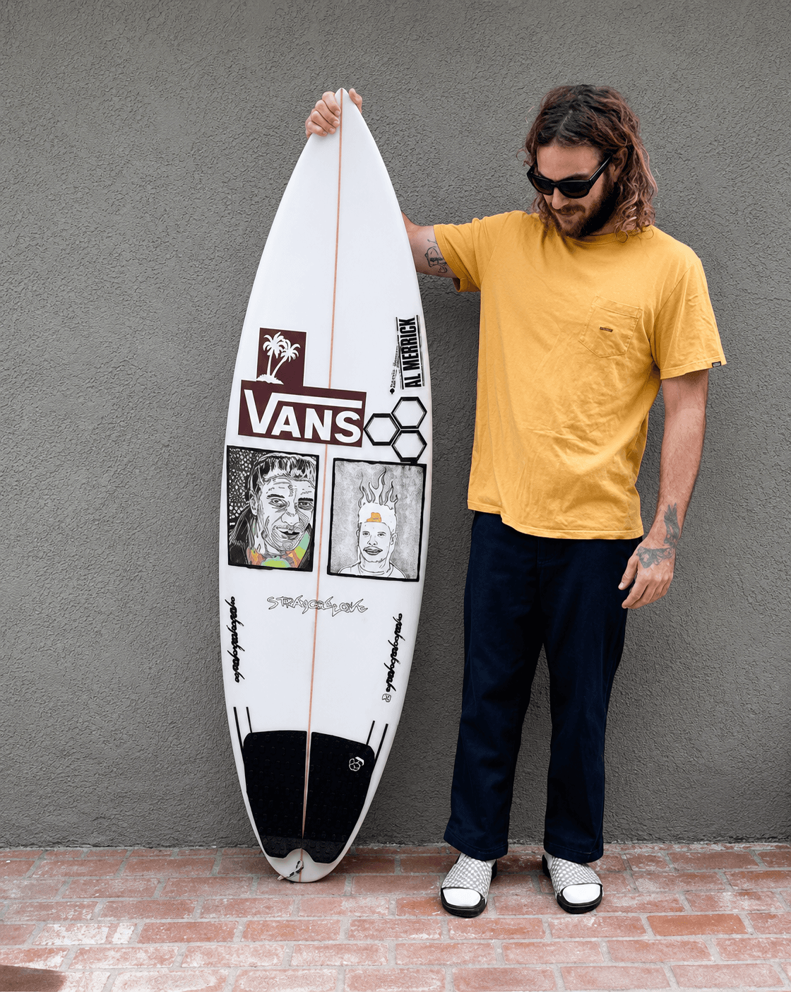 A man with a beard and sunglasses holds a white surfboard adorned with Vans stickers and graphic artwork against a gray wall.